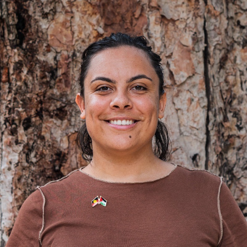 Portrait of woman wearing a brown tshirt in front of a tree. the woman is smiling. she wears a pin with the Aboriginal and Palestinian flags.